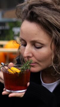 Woman examines herb garnished cocktail at outdoor cafe, bringing its glass near her face to smell drink. Relaxed customer appears thoughtful while appreciating its aroma before tasting cocktail.