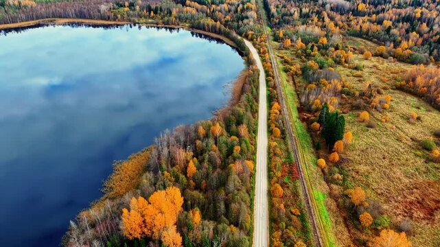 Colorful autumn forest landscape reflecting on still lake near winding road and railway tracks