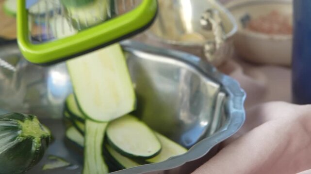 Hands slicing zucchini over a steel bowl with a mandoline slicer