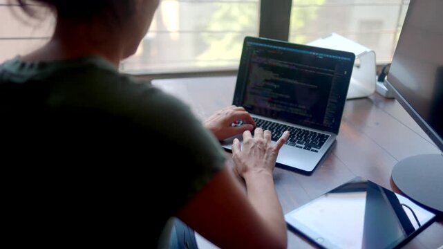 Unrecognizable female programmer coding on laptop computer sitting at her desk by the window, focused on writing html code, developing new software application or website