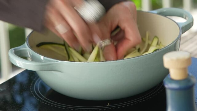 Hands adding zucchini strips to shrimp in a pale blue pan