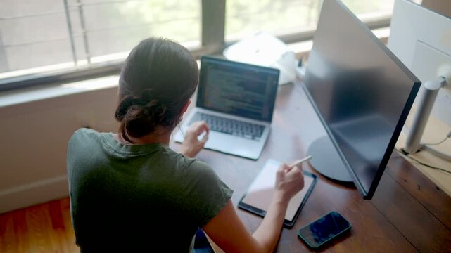 Woman software developer coding on a laptop and writing on a digital tablet, multitasking at her modern home office desk with multiple devices and a computer monitor