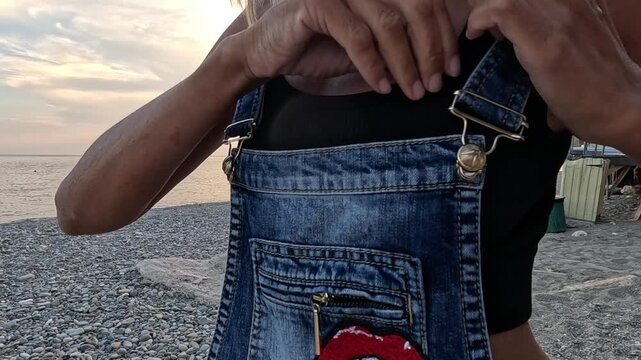 Denim overalls adjustment by woman standing on coastal shingle beach during dusk. Medium closeup shot.
