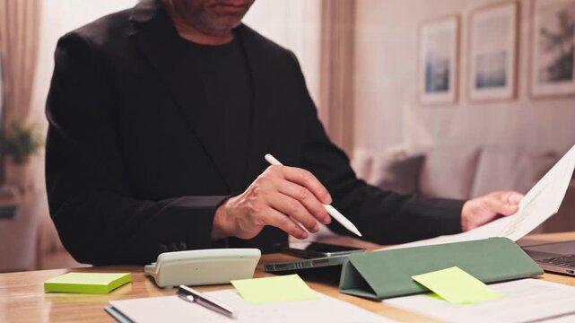 Mature businessman reviews financial papers holds electronic pen sitting desk home office. Corporate tax calculation, investment portfolio management, financial audit analysis paperwork review.