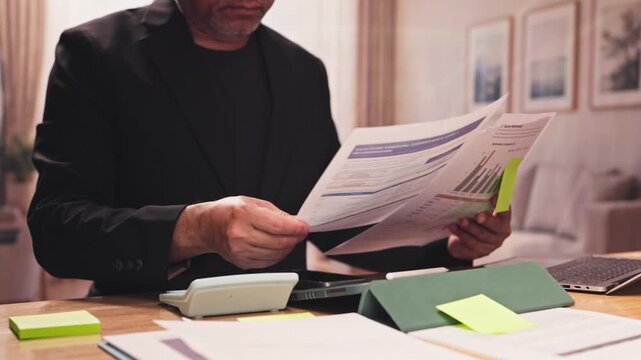 Mature businessman calculates financial data sheets, charts, invoices seated indoor. Corporate budget planning, investment portfolio audit, annual revenue accounting review.