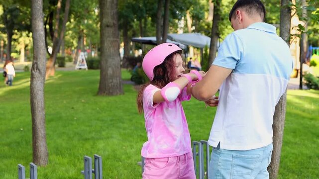 Father comforting his upset crying daughter while helping her adjust protective gear in a summer park in slow motion. Active kids safety and parenting concept.