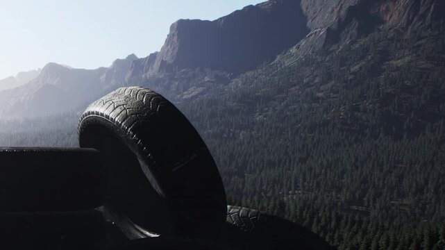 Large black rubber tires stand against a backdrop of deep green evergreen trees covering steep mountain slopes during the bright morning light as fog lingers in the distance across the rocky terrain.