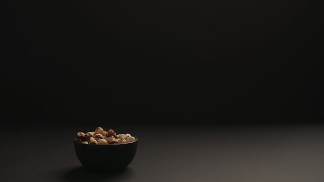 Slow motion roasted hazelnuts fall into black bowl on black paper background