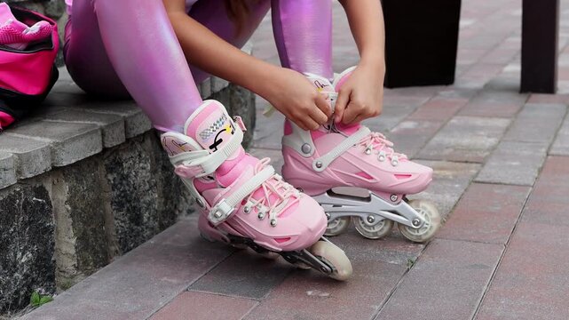 Close-up of a little girl in a pink helmet and protective gear fastening straps on her inline roller skates while sitting on stone steps outdoors. Rollerblading and active kid leisure concept.