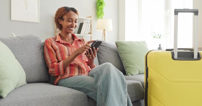 Woman booking vacation travel on smartphone. Happy traveler uses a mobile device near yellow luggage, arranging accommodation for holiday journey from home. Easy online trip planning concept.