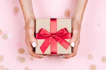 Female hands with milky manicure holding a kraft gift box with red satin bow on a pink background with pastel confetti. © Anna Efetova