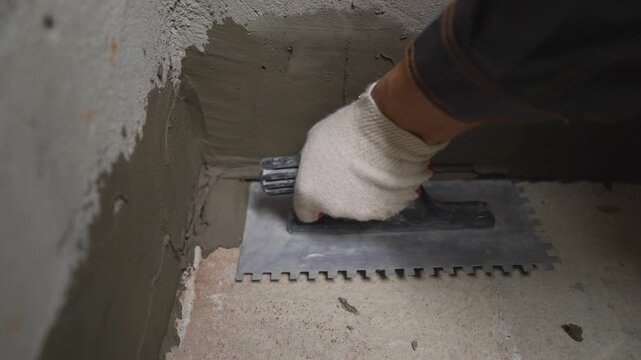 Close up of a construction worker in gloves using a notched trowel to apply wet cement or plaster to a concrete corner. This shows a waterproofing or tiling process during home renovation or repair