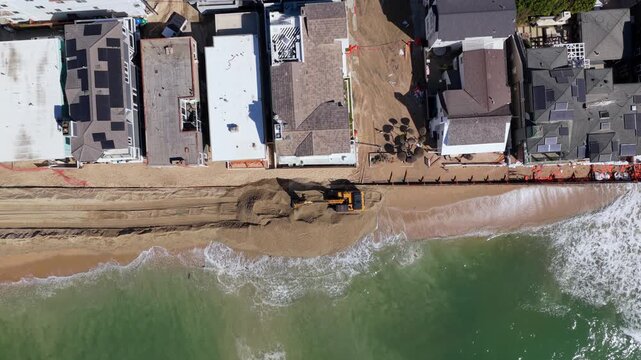 Long Beach, California, USA - October 15, 2026: Static top down view of an excavator piling sand into a flood berm as waves wash up to the broken seawall below homes after Tropical Storm Rachel.