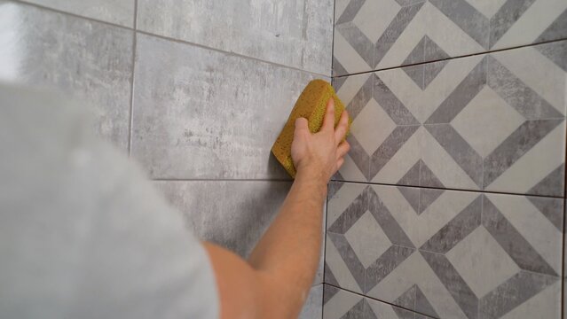 Worker's hand with yellow sponge cleaning excess grout from new grey and white geometric ceramic wall tiles, home improvement project