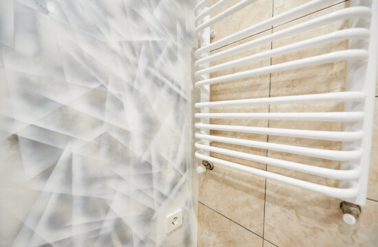Close up of heated towel rail on beige bathroom tiles beside abstract accent wall. Modern washroom detail with clean lines, minimalist decor and contemporary interior design