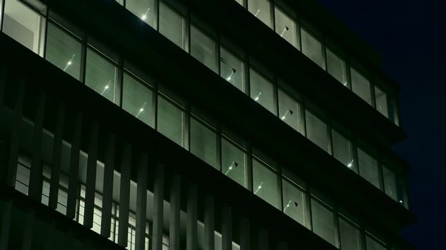 A low angle view shows stacked floors and glowing windows on a modern office building against the night sky in Tokyo Japan. This highlights the scale and geometry of urban architecture.