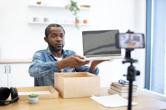 An African American man presents a laptop to a smartphone camera, with cardboard packaging, books, and desk accessories in a bright workspace
