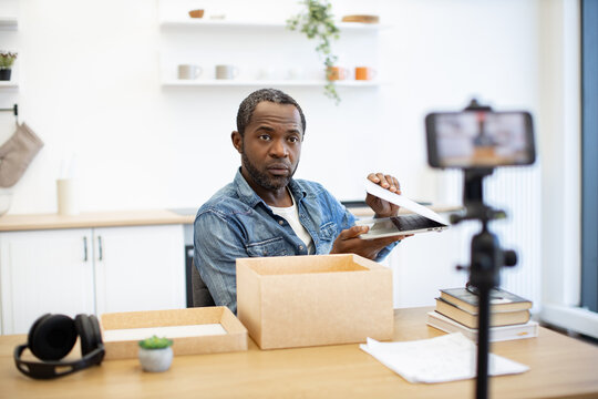 A content creator records a laptop unboxing at a bright home workspace, with cardboard packaging, books, headphones, and a phone mounted on a tripod