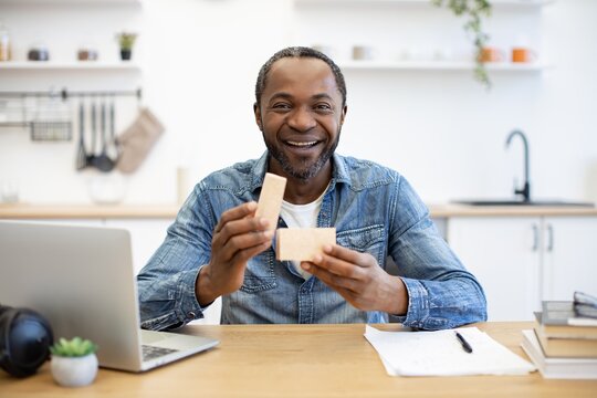 A smiling African American man holds two small cardboard pieces at a wooden desk, with a laptop, notebook, and pen in a bright home workspace