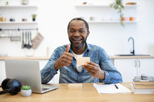 Working at a wooden table, he holds a cup and gives a thumbs-up beside a laptop, notebooks, and headphones in a bright home kitchen interior