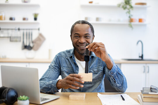 A smiling African American man examines a small device at his home workspace, with a laptop, headphones, notebook, and bright kitchen in the background