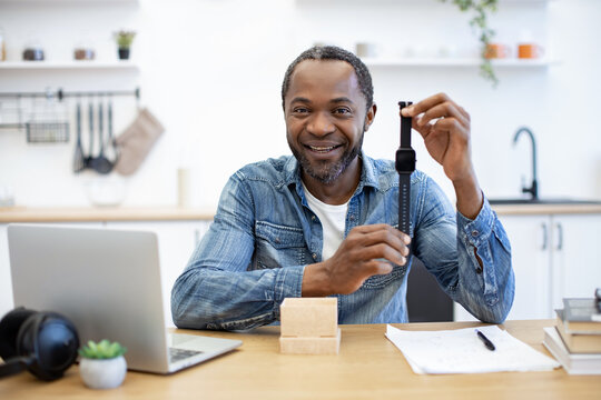 A smiling African American man examines a handheld measuring tool at a desk, with laptop, notes, and headphones in a bright home workspace