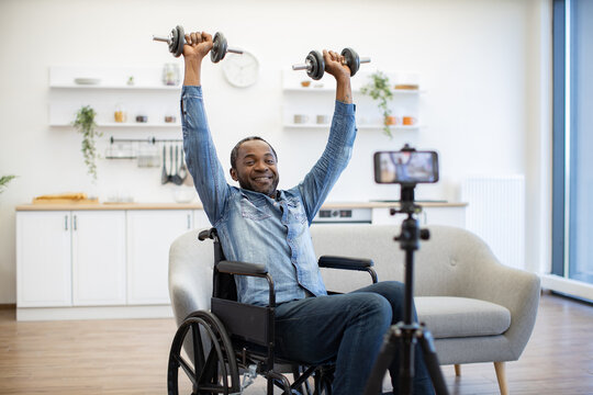 A smiling man lifts dumbbells overhead from his wheelchair in a bright home, while a smartphone mounted on a tripod faces him during his workout