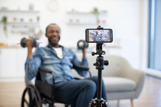 A man in a wheelchair lifts dumbbells while recording a home fitness session on a smartphone mounted to a tripod in a bright living room