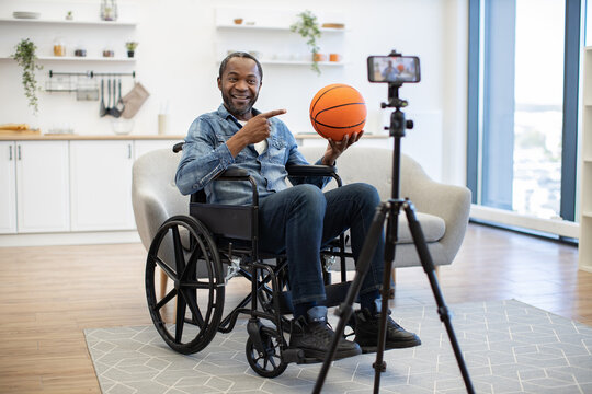 A smiling African American man in a wheelchair holds up a basketball and points toward a smartphone camera in a bright, modern home interior