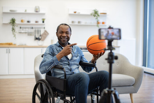 A smiling African American man in a wheelchair holds a basketball and gestures toward a smartphone recording him in a bright, modern home interior