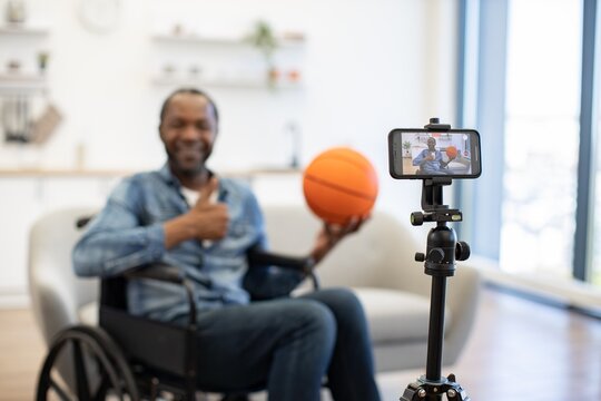 A wheelchair user records a video at home while holding an orange basketball, with a smartphone mounted on a tripod visible in the foreground