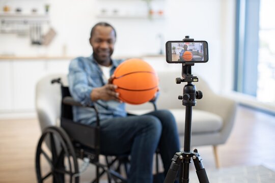 A wheelchair user holds an orange basketball while a smartphone on a tripod records him in a bright, modern living room with soft natural light