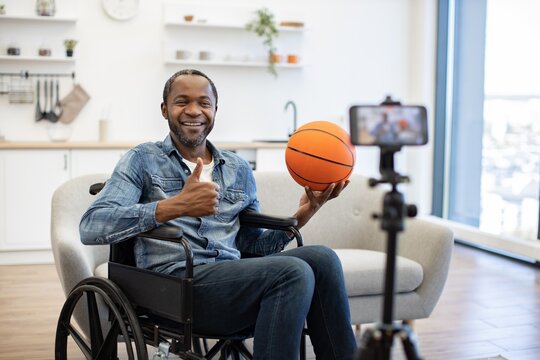 A smiling man in a wheelchair gives a thumbs-up while holding a basketball, recording a cheerful video in a bright, modern home interior with a phone on a tripod nearby