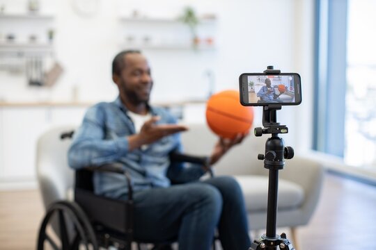 A wheelchair user holds a basketball while speaking to a smartphone on a tripod in a bright home, creating accessible sports content indoors