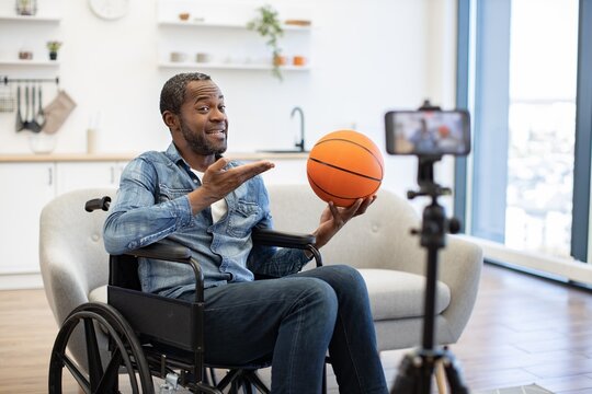 A seated man gestures toward an orange basketball at home as a smartphone mounted on a tripod captures the moment in a bright, open living space
