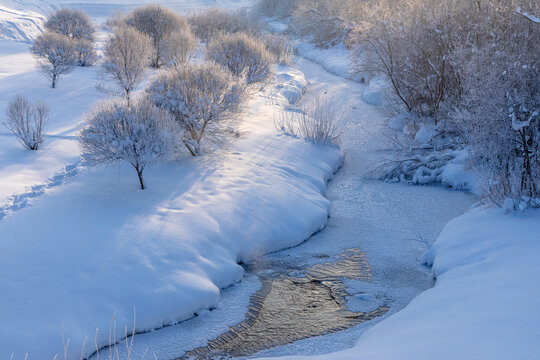 Winding frozen river with icy patches between deep snow banks and hoarfrost-covered bushes and trees under soft winter sunlight, serene landscape