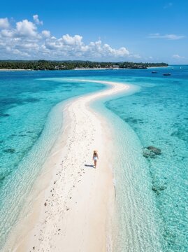 A person walks along a pristine, white sand beach that stretches out into a turquoise ocean. The beach is a narrow, winding path leading to a distant island.