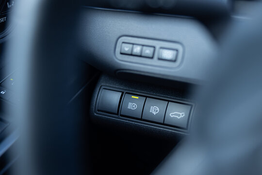 Close view of vehicle steering column controls with illuminated buttons, headlight adjustment, and trunk release switches in a modern automobile interior dashboard panel