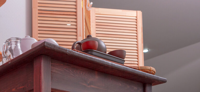Tea set with teapot, cups and glass pitcher on wooden counter beneath louvered shutters. Tea service and traditional brewing utensils. Home kitchen, hospitality and beverage preparation