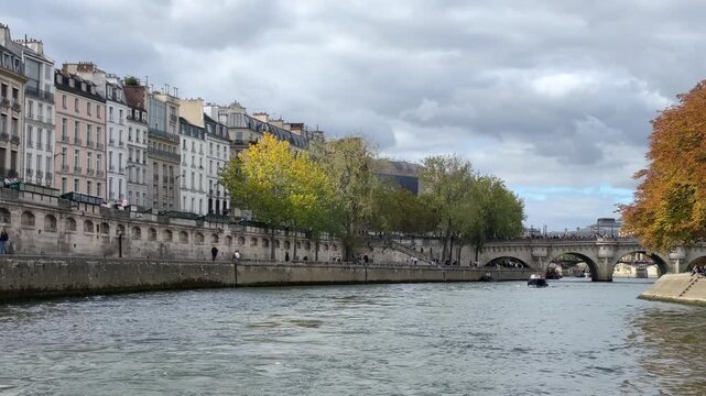 View from a tour boat cruising the Seine River in Paris, France, passing historic buildings and autumn trees as protesters cross a stone bridge in the distance. 15450  