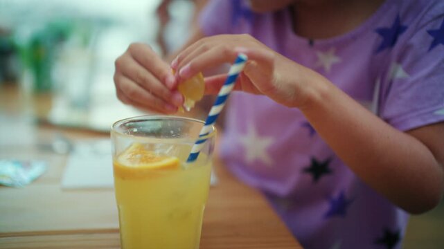 Close-up of a girl squeezing an orange slice into her iced drink with a blue striped paper straw, French Riviera, France.