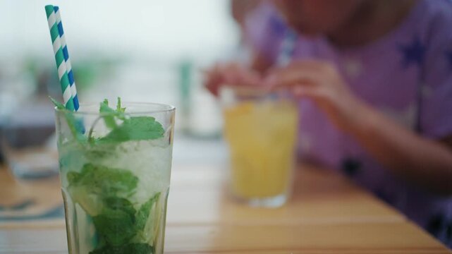 Close-up of a mojito with fresh mint, ice and a striped straw, a girl with an orange drink out of focus behind, French Riviera, France.