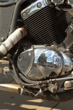 Close-up of motorcycle engine and exhaust components showing chrome cover, cooling fins, wrapped exhaust pipe and linkage hardware.