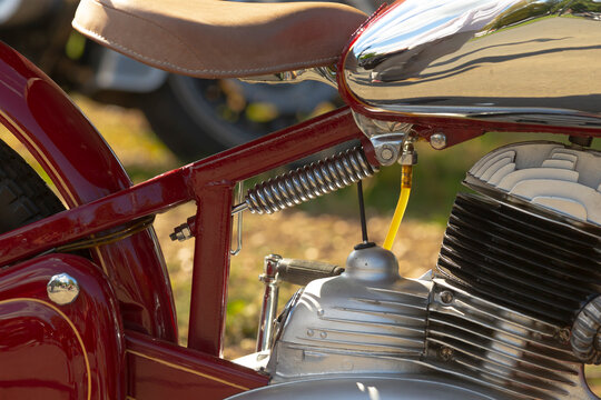 Close-up of motorcycle rear frame showing leather seat, chrome fuel tank, coil spring suspension, yellow fuel line, and engine cooling fins in natural daylight.