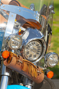 Close-up view of motorcycle front showing chrome headlamp cluster, auxiliary lights, windshield hardware, and leather fringe tool roll attached to fork.