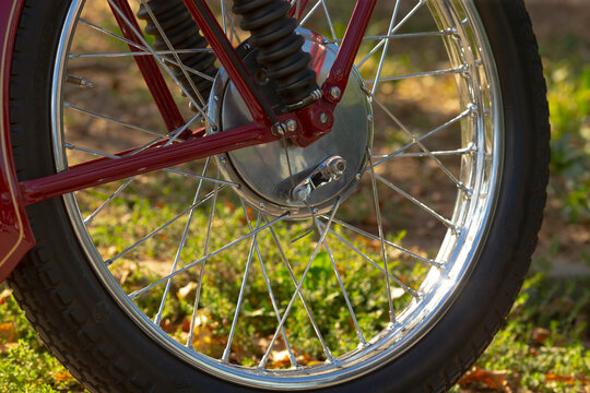 Close-up of motorcycle front wheel showing chrome spokes, metal hub, tire tread, and red-painted fork against grass background.