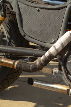 Close-up of motorcycle exhaust pipe and wrapped header with adjacent rear tire and metal side panel showing diamond plate texture.