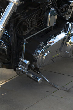 Close-up view of chrome engine casing, footpeg, and kickstand on parked motorcycle captured on paved sidewalk surface with reflections on metal parts.