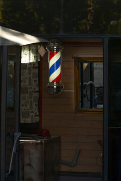 Interior view of small barbershop showing illuminated red white blue striped barber pole mounted on wooden wall next to framed mirror and patterned curtain.