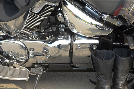 Close-up of chrome engine casing and footrest on parked motorcycle with pair of black leather riding boots beside it reflecting surrounding pavement and figures.
